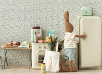 A stuffed bunny in a striped shirt and blue shorts opens a retro-style fridge in a tiny kitchen with the Maileg Grocery Box, Miniature, tote bag with carrot, Maileg Mouse accessories, and floral wallpaper—perfect for imaginative play.