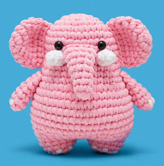 Lulu the Elephant Crochet Kit by The Woobles features a pink stuffed elephant with big ears, a long trunk, black button eyes, and white accents, set against a bright blue background. Ideal for beginner crocheters or as a gift.