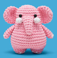 Lulu the Elephant Crochet Kit by The Woobles features a pink stuffed elephant with big ears, a long trunk, black button eyes, and white accents, set against a bright blue background. Ideal for beginner crocheters or as a gift.