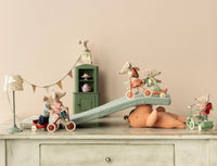 Four toy mice ride scooters and a Maileg Tricycle Hanger, Mouse - Coral by Maileg on a soft ramp over a large stuffed bear, with mouse accessories, a vintage cabinet, lamp, and colorful bunting in the background.