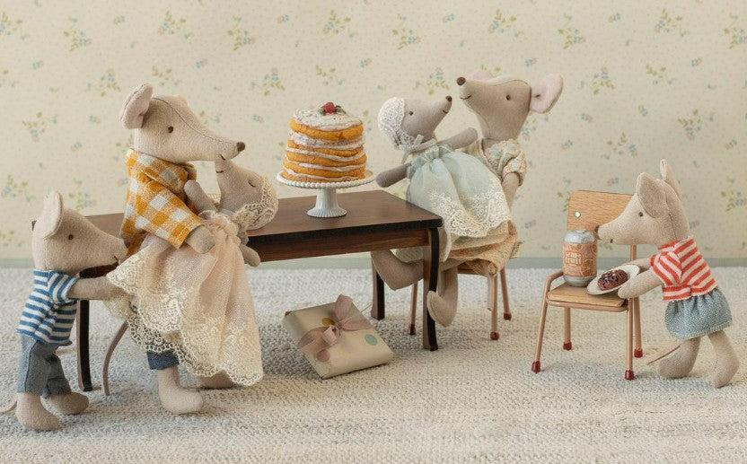 Five dressed Maileg mice gather around a table with the Maileg Party Cake And Cake Stand, Mouse - Off White. One mouse sits with a bottle, two hold hands, another hugs a small mouse, while accessories and a wrapped gift lie on the floor.