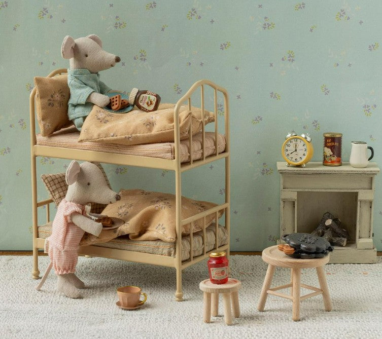 Two toy mice in pajamas sit on the Maileg Bunk bed, Mouse - Wheat by Maileg, each holding a small book. The scene is styled with delightful mouse-house furniture—including a nightstand with a clock, mugs, and stove, plus stools and miniature food items—creating a cozy, vintage-inspired dollhouse setting.