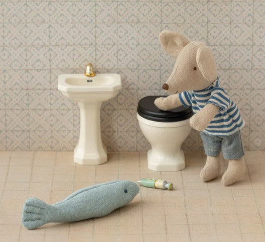 A Maileg mouse in a striped shirt stands beside the Maileg Bathroom sink, Mouse - Off white, gazing at a soft toy fish and a small tube on the tiled floor of its charming miniature bathroom.