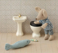 A Maileg mouse in a striped shirt stands beside the Maileg Bathroom sink, Mouse - Off white, gazing at a soft toy fish and a small tube on the tiled floor of its charming miniature bathroom.
