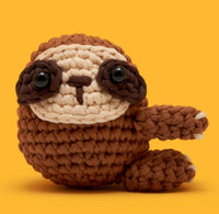 Sam the Sloth Crochet Kit by The Woobles features a brown and cream plush sloth with large black eyes and a raised arm, posed against a yellow background—the ideal beginner crochet project.