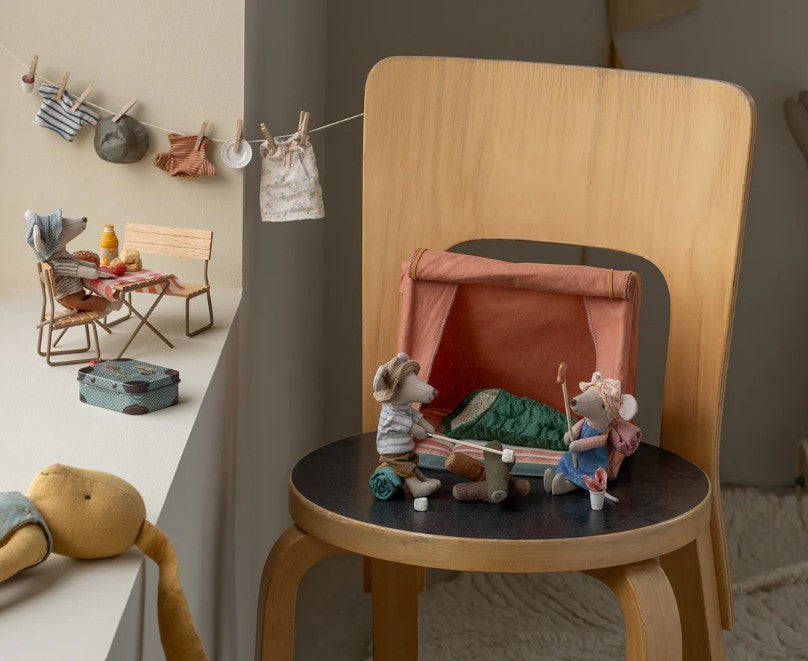 Maileg Hiker Mouse, Big Sister by Maileg sits at a miniature tent on a wooden chair, ready for camping fun. In the background, more Maileg mice picnic and tiny clothes hang nearby, creating a playful indoor scene inspired by outdoor adventures.