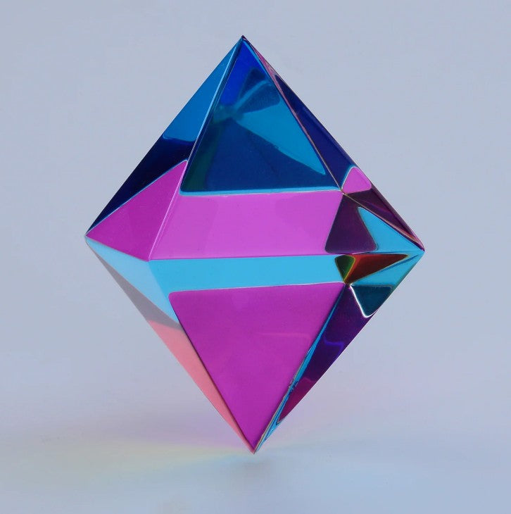 The Aether by CMY Cubes is a geometric, diamond-shaped optical cube with reflective pink, blue, and purple surfaces on a plain background—ideal as a sensory display for stress relief.