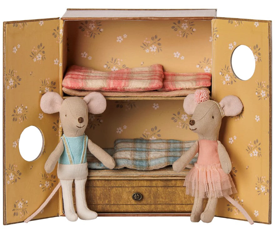 Two Maileg Ballet mice—Little Sister and Brother—stand before a flower-adorned yellow box that transforms into their cozy miniature dance studio. Dressed for ballet-themed adventures, the brother mouse sports a blue outfit while Little Sister wears a pink dress with a floral headband. Inside, plaid-blanketed bunk beds await, inviting endless imaginative play from this charming Maileg set.