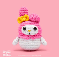A plush white rabbit with a pink hood, yellow bow, and black features—made using the Woobles My Melody Crochet Kit—is displayed on a solid pink background.