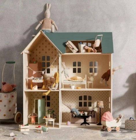 The Maileg House of Miniature Dollhouse by Maileg is a detailed playhouse featuring animal dolls, miniature furniture, and charming toys. A plush rabbit doll sits atop the roof, creating a playful scene that evokes a whimsical miniature world. Nearby on the neutral background are a stroller and storage basket, completing the delightful setup.