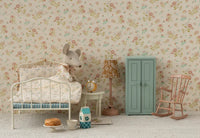 A cozy, vintage-style kids’ room features a Maileg Alarm Clock, Mouse - Yellow by Maileg on the bedside table, a white metal bed with floral bedding, mouse plush toy, mint green wardrobe, pink rocking chair, and toy food on a floral wallpapered floor.