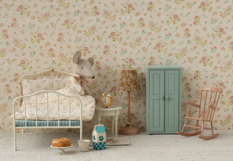 A cozy, vintage-style kids’ room features a Maileg Alarm Clock, Mouse - Yellow by Maileg on the bedside table, a white metal bed with floral bedding, mouse plush toy, mint green wardrobe, pink rocking chair, and toy food on a floral wallpapered floor.