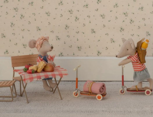 Two Maileg mice enjoy imaginative indoor play: one sits at a table with delicious treats, while the other stands on the Maileg Kick board, Mouse - Orange, wearing a tiny backpack—all set against an enchanting floral wallpaper backdrop for endless make-believe adventures.