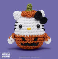 The Woobles Halloween Hello Kitty Crochet Kit features a plush Hello Kitty dressed as a glittery orange jack-o-lantern with a green stem, set against a purple background—perfect for any Hello Kitty crochet collection.