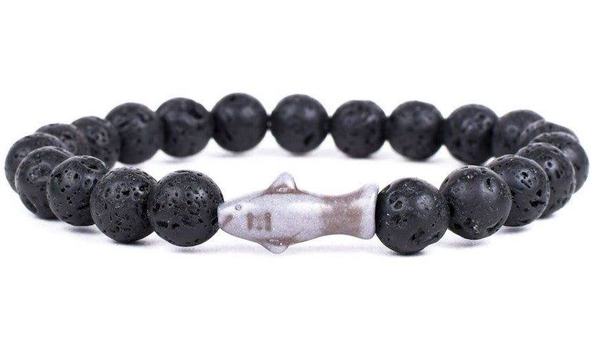 The fahlo Voyage - Lava Stone Shark bracelet showcases round black lava stones with a central white shark-shaped bead—ideal for ocean conservation enthusiasts.