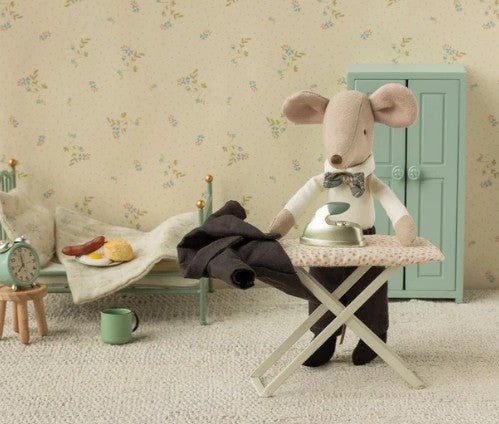 A Maileg mouse in a bow tie stands at the Maileg Iron and ironing board, Mouse, carefully ironing tiny pants in a whimsical dollhouse room featuring a green bed, cupboard, cup, alarm clock, and a plate of food. This charming setting is enhanced with delightful Maileg mice accessories and the adorable miniature ironing set by Maileg.