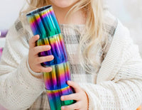 A young child with long blonde hair holds ahimsa's Rainbow Conscious Cups (8 oz, pack of 4) while wearing a plaid dress and a cream-colored knitted cardigan.