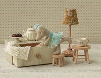 The Maileg Dance Mouse In Daybed, Little Sister by Maileg lounges in a striped box with miniature furniture—lamp, cake, stools, and daybed—inviting imaginative play against a charming patterned background.