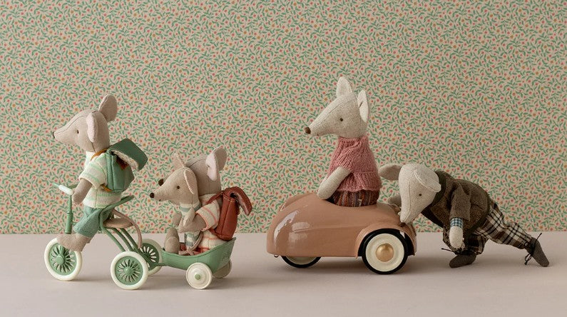 Four plush Maileg mice play with vintage-style toys—two ride the green Maileg Tricycle Garage, one sits in a pink car, and another pushes it. A small floral background complements Maileg mouse accessories or miniature storage.