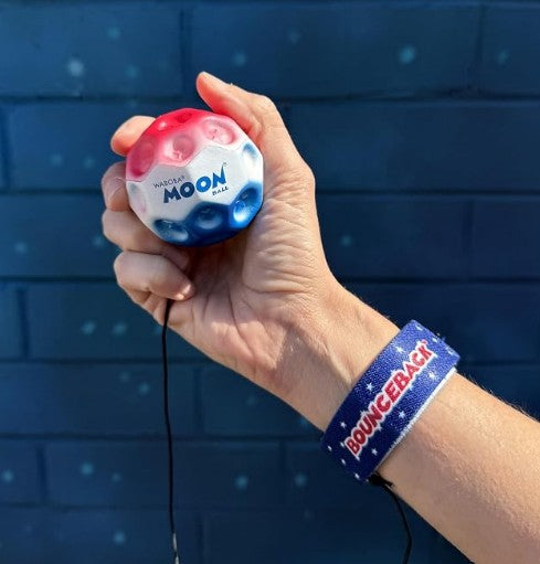 A hand holds a Waboba USA / Stars & Stripes Bounceback ball adorned in red, white, and blue. The person wears a bold blue wristband inscribed with BOUNCEBACK. Set against a vivid blue-painted brick wall, the scene perfectly captures the spirit of outdoor active play.