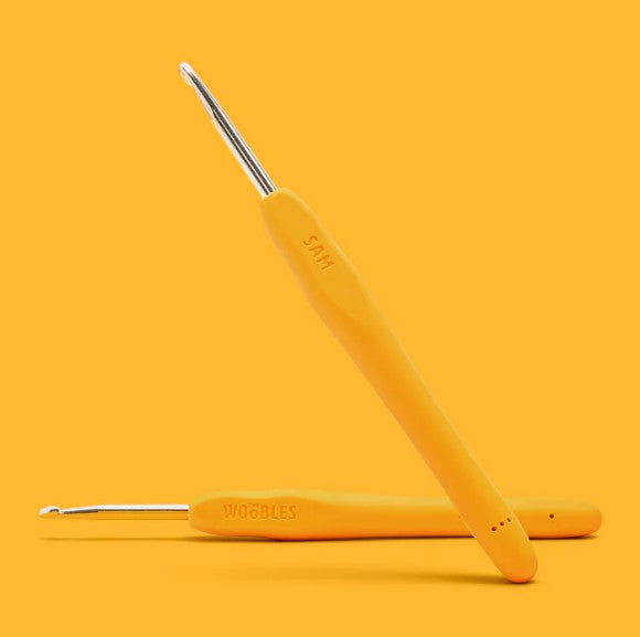 Two yellow ergonomic crochet hooks from The Woobles’ Sam the Sloth Crochet Kit are displayed on a yellow background. One hook is flat and the other leans, with "WOBBLES" and "4MM" visible on their handles.