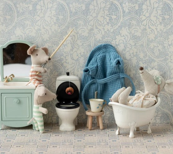 Three toy mice in a bathroom inspire imaginative play: one fishes in a toilet, one relaxes in a Maileg miniature bathtub, and another stands by a vanity with a mirror and drawer. The Maileg Bathrobe, Dad mouse - Dusty blue from Maileg hangs on the wall, while a small stool holds a cup.