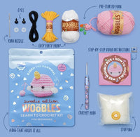 A beginner-friendly crochet kit by The Woobles, “Bjorn the Narwhal: Sweetie Edition,” is shown on a blue background with yarn, eyes, needle, stuffing, hook, video instructions, and a bag featuring a pink narwhal plush.