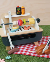 The Tenderleaf Barbeque Play Set features wooden skewered veggies, steak, and sausages on a red checkered picnic blanket, with pretend BBQ sauce and mustard bottles as fun play food accessories.
