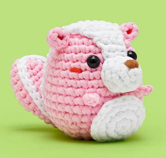The Woobles Andre the Skunk Pink Crochet Kit is a beginner-friendly set to make a plush skunk toy with pink and white yarn, black button eyes, a small brown nose, and a fluffy tail, all set against a light green background.