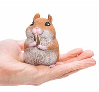 A realistic, small Chonky Cheeks Hamster figurine by Schylling Toys, featuring brown-and-white coloring and holding a sunflower seed, sits upright in the palm of a person's hand against a white background—making it an adorable sensory toy that's perfect for stress relief.