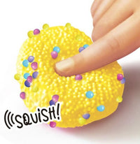 A hand presses a finger into a yellow, textured Craze Sensations ASMR fidget toy from License 2 Play, filled with colorful beads. The word SQUISH! highlights the soft, squeezable texture and sensory fun of this playful collectible.