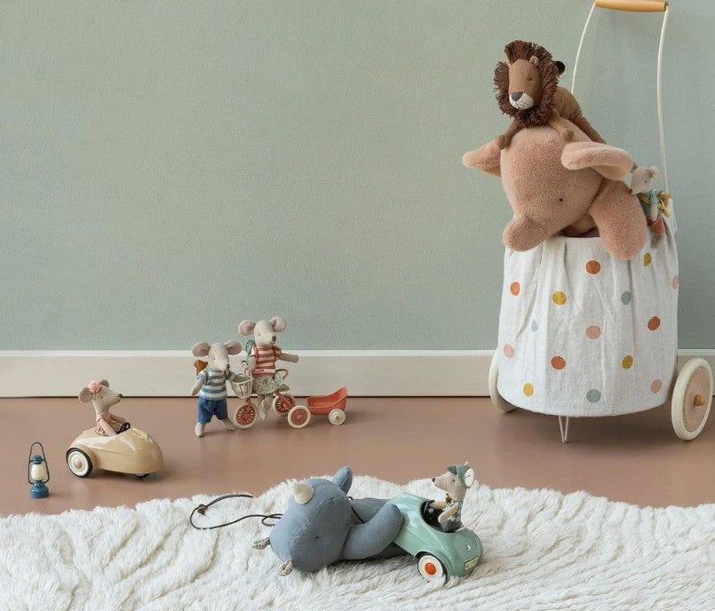 Several Maileg mice toys ride in a Maileg Car, Mouse – Light mint on a light brown floor near a white rug, while a stuffed lion sits atop a polka-dotted basket with wheels against a light green wall.