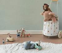 Several Maileg mice toys ride in a Maileg Car, Mouse – Light mint on a light brown floor near a white rug, while a stuffed lion sits atop a polka-dotted basket with wheels against a light green wall.