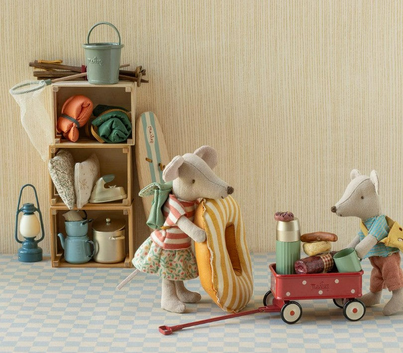 Two Maileg mice, geared up for camping, stand beside the Maileg Wagon, Mouse - Red by Maileg, which is loaded with food and drinks and ready for their next tiny adventure. Behind them, a shelf displays camping supplies—pots, a lantern, and rolled sleeping bags—set against a beige wall and checkered floor.