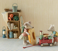 Two Maileg mice, geared up for camping, stand beside the Maileg Wagon, Mouse - Red by Maileg, which is loaded with food and drinks and ready for their next tiny adventure. Behind them, a shelf displays camping supplies—pots, a lantern, and rolled sleeping bags—set against a beige wall and checkered floor.