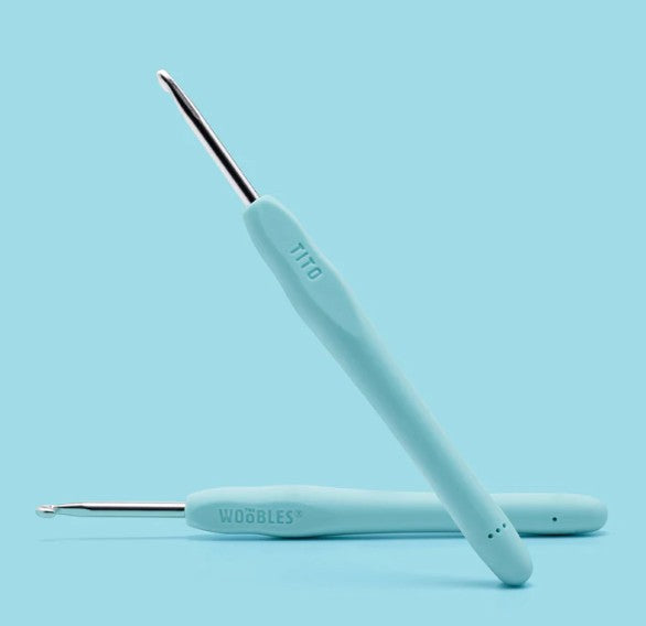 Two light blue ergonomic crochet hooks from The Woobles’ Tito the Swan Pastel Crochet Kit are displayed on a matching background. Handles labeled TITO and WOOBLES; one hook lies flat, the other stands at an angle—ideal for beginners.