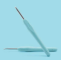 Two light blue ergonomic crochet hooks from The Woobles’ Tito the Swan Pastel Crochet Kit are displayed on a matching background. Handles labeled TITO and WOOBLES; one hook lies flat, the other stands at an angle—ideal for beginners.