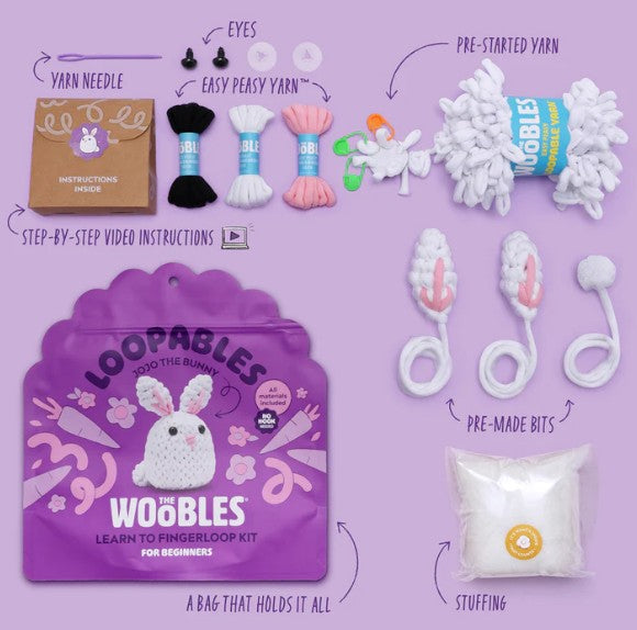 The Woobles Jojo the Bunny Loopables Kit is displayed on a purple surface, featuring yarn, a yarn needle, instructions, eyes, bunny ears, stuffing, pre-made pieces, a storage bag, and pre-started yarn to create a white bunny.