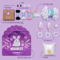 The Woobles Jojo the Bunny Loopables Kit is displayed on a purple surface, featuring yarn, a yarn needle, instructions, eyes, bunny ears, stuffing, pre-made pieces, a storage bag, and pre-started yarn to create a white bunny.