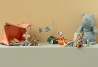 A delightful Maileg display: mice with the Maileg Bonfire Set, one in a tent, another rowing an orange raft, one fishing by the bonfire set, plus a blue stuffed rhino—all with charming mini props on a beige background.