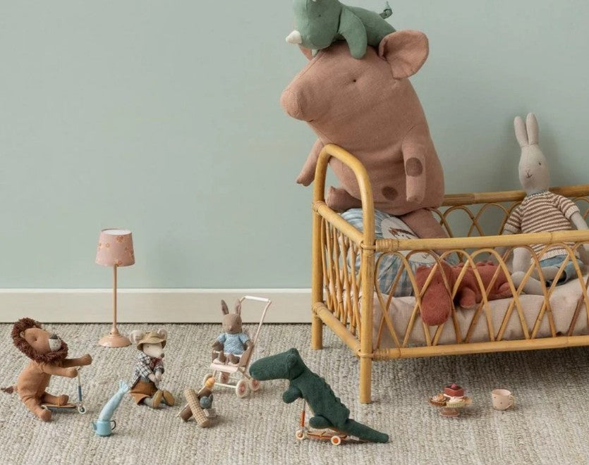 A cozy nursery features a rattan crib with plush toys and Maileg mice. On the floor, the Maileg Bonfire Set, Mouse by Maileg, tea set, camping accessories, a lamp, and a stroller sit against a pale green wall.