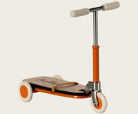 The Maileg Kick Board, Mouse - Orange by Maileg is a small, vintage-style orange scooter with white handlebars and a wooden deck—perfect for Maileg Mice—with one front wheel and two rear wheels featuring white tires.