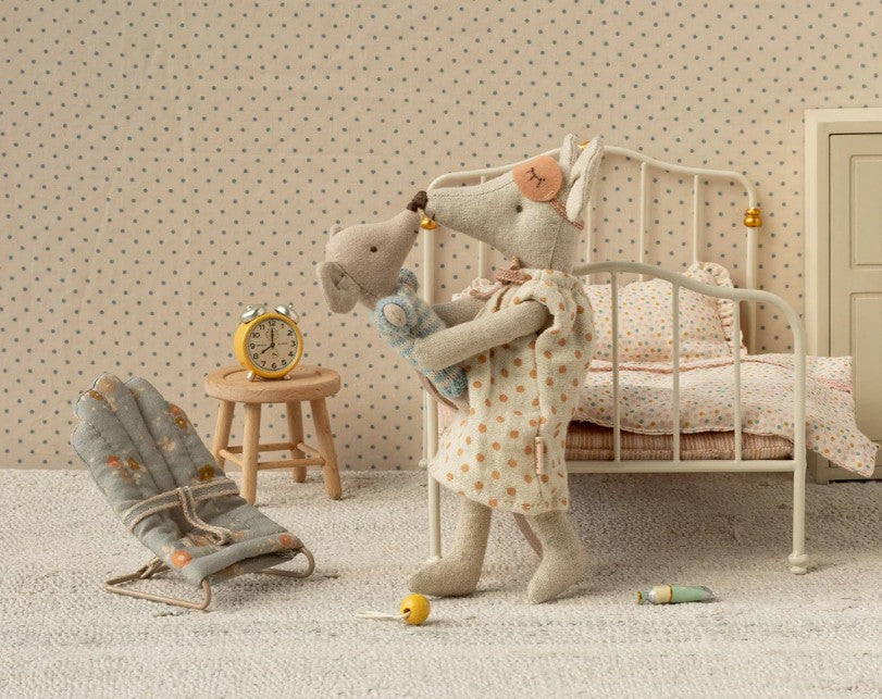 Two Maileg My-sized fabric mice—one adult in a nightgown and one child in pajamas—hug beside a small bed. Nearby are a stool with yellow alarm clock, mini seat, scattered sewing items, and the Maileg Babysitter, My - Blue.