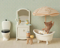 A plush Maileg mouse relaxes in a white toy bathtub under the Maileg Beach Umbrella, Mouse - Soft Coral, surrounded by miniature bathroom furniture—a toilet, sink, mirror, and stool with bath items—set against patterned wallpaper.
