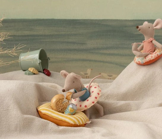 Two Maileg toy mice with swim rings relax on a sandy beach scene crafted from fabric, while a Small Mouse enjoys pretend play nearby with a green bucket, seashells, and the Maileg Rubber boat, Small mouse - Yellow stripe floating in the calm water in the background.