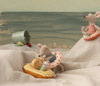 Two Maileg toy mice with swim rings relax on a sandy beach scene crafted from fabric, while a Small Mouse enjoys pretend play nearby with a green bucket, seashells, and the Maileg Rubber boat, Small mouse - Yellow stripe floating in the calm water in the background.