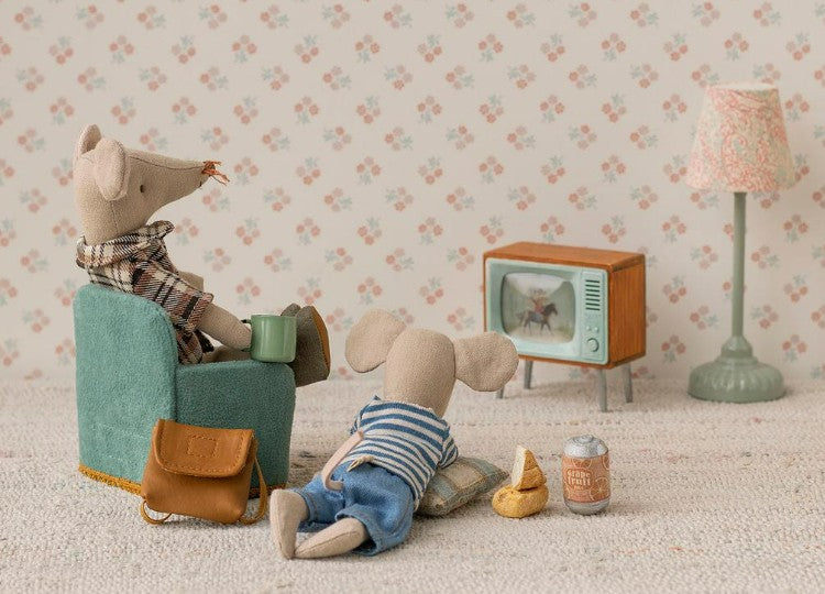 Two Maileg Mouse toys enjoy a cozy room with the Maileg Television, Mouse by Maileg. One mouse sits in an armchair with a mug, the other lounges on the floor. Snacks, a lamp, and floral wallpaper create a charming dollhouse scene.