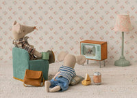 Two Maileg Mouse toys enjoy a cozy room with the Maileg Television, Mouse by Maileg. One mouse sits in an armchair with a mug, the other lounges on the floor. Snacks, a lamp, and floral wallpaper create a charming dollhouse scene.