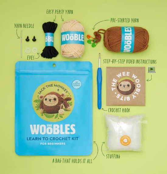 Flat lay of The Woobles Jack the Monkey Crochet Kit, including yarn bundles, a blue crochet hook, yarn needle, eyes, stuffing, step-by-step instructions, and a blue storage bag on a green background.