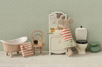Maileg Tricycle Mouse, Big Sister - Green by Maileg features Big Sister in a striped top and floral skirt with a tricycle, purse, and bathroom set—complete with vanity, bathtub, toilet, chair, towel—all on a textured floor for imaginative play.